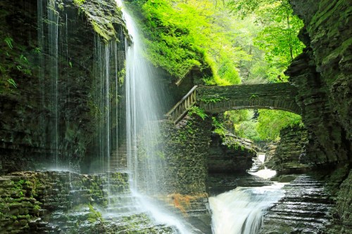 A serene waterfall cascades over moss-covered rocks, flowing beneath a stone bridge in a lush, green canyon.
