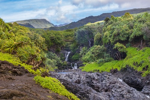Lush green landscape with waterfalls cascading into a rocky stream, surrounded by mountains and a blue sky.