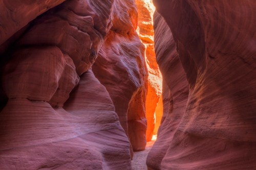 A narrow canyon with smooth, reddish walls and a bright light shining at the end, creating a warm glow.