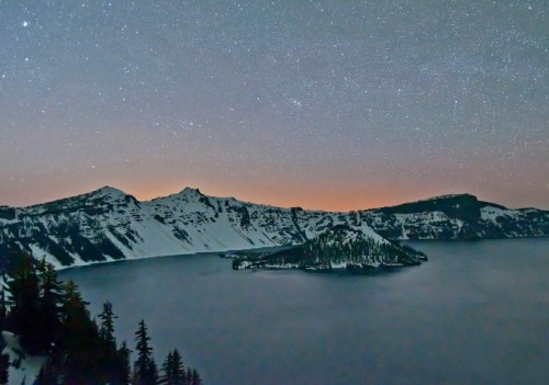 A starry night sky over a snow-covered mountain lake, with an island in the center and silhouettes of trees in the foreground.