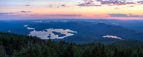 A panoramic view of mountains and lakes at sunset, with vibrant colors reflecting on the water and a forest in the foreground.