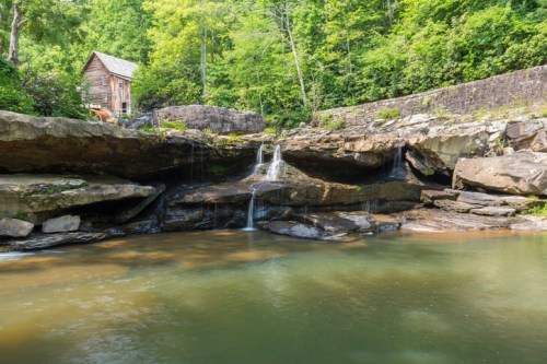 A serene landscape featuring a small wooden cabin beside a rocky stream and lush green trees.
