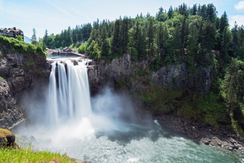 A stunning waterfall cascades into a river, surrounded by lush greenery and rocky cliffs under a clear blue sky.
