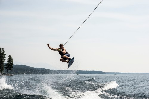 A person jumps off a wakeboard, soaring above the water with a rope in hand, surrounded by trees and a calm lake.