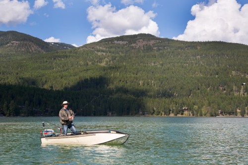 A person fishing from a small boat on a calm lake, surrounded by lush green mountains and a blue sky with clouds.