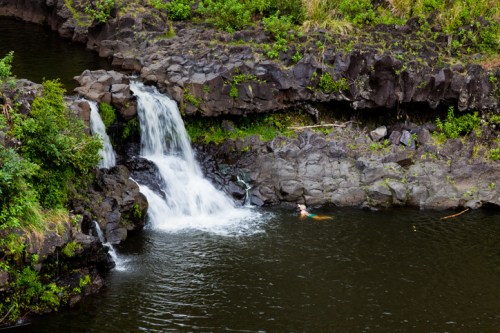 A serene waterfall cascades into a dark pool surrounded by rocky terrain and lush greenery.