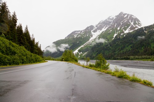 A winding road alongside a river, with lush green trees and a snow-capped mountain in the background under cloudy skies.