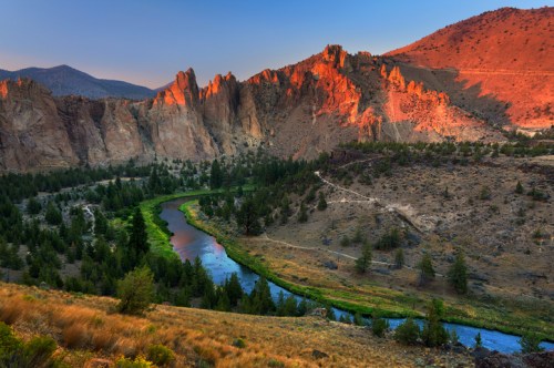 A winding river flows through a rugged canyon, surrounded by rocky cliffs and lush greenery under a colorful sunset.