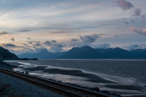 A serene landscape featuring mountains, a calm sea, and a railway track along the shore at dusk.