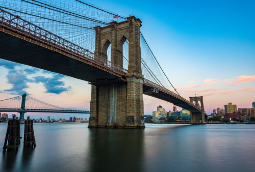 Brooklyn Bridge at sunset, with the Manhattan skyline and another bridge in the background, reflecting on the water.