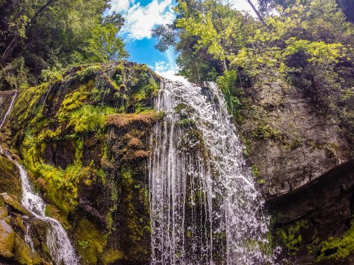 A lush waterfall cascading over moss-covered rocks, surrounded by vibrant green trees and a bright blue sky.