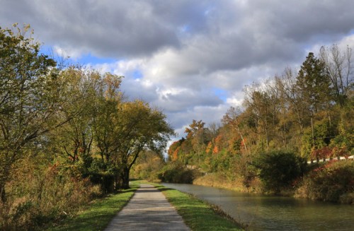 A scenic pathway lined with trees, alongside a calm waterway, under a cloudy sky with hints of autumn colors.