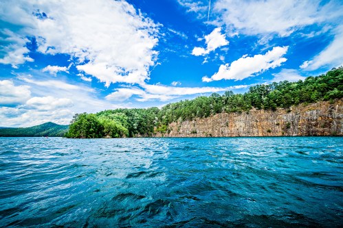 A serene lake surrounded by lush greenery and rocky cliffs under a bright blue sky with fluffy clouds.