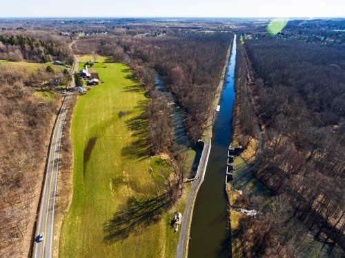 Aerial view of a winding river alongside a green field and a road, with trees lining the banks in a rural landscape.