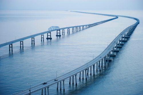 A long, winding bridge stretches over calm waters, connecting land with a distant horizon under a soft blue sky.