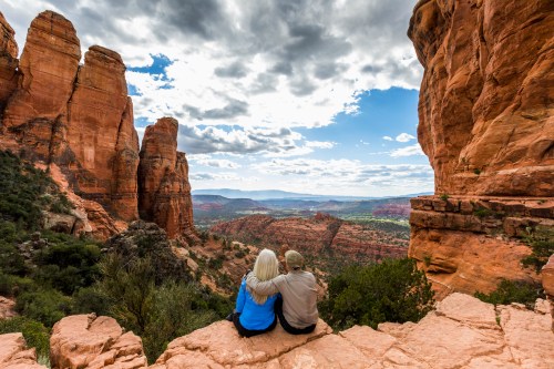 A couple sits on a rock ledge, overlooking a scenic valley with red rock formations and a cloudy sky.