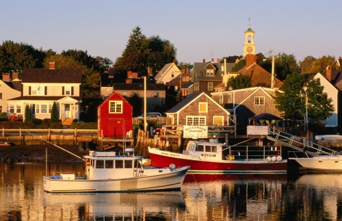 A serene harbor scene with boats, colorful houses, and a clock tower reflecting in calm waters at sunset.