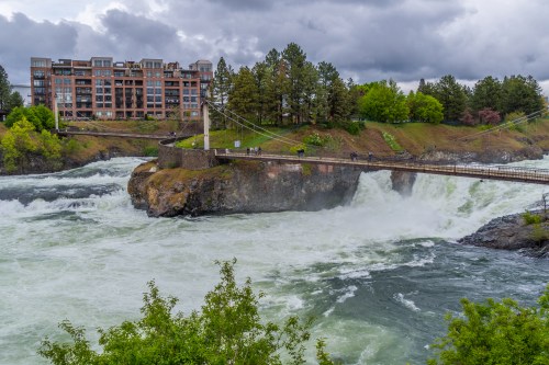 A suspension bridge over a rushing river, with a modern building and lush trees in the background under a cloudy sky.
