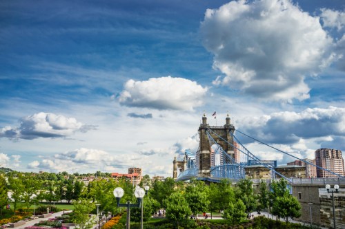 A scenic view of a bridge with blue towers, surrounded by greenery and a bright sky with fluffy clouds.