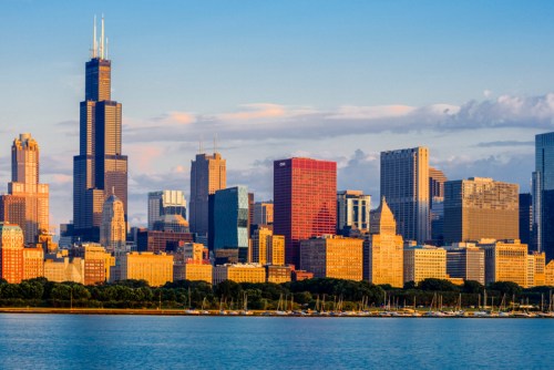 Chicago skyline at sunset, featuring tall buildings and a calm lake in the foreground.