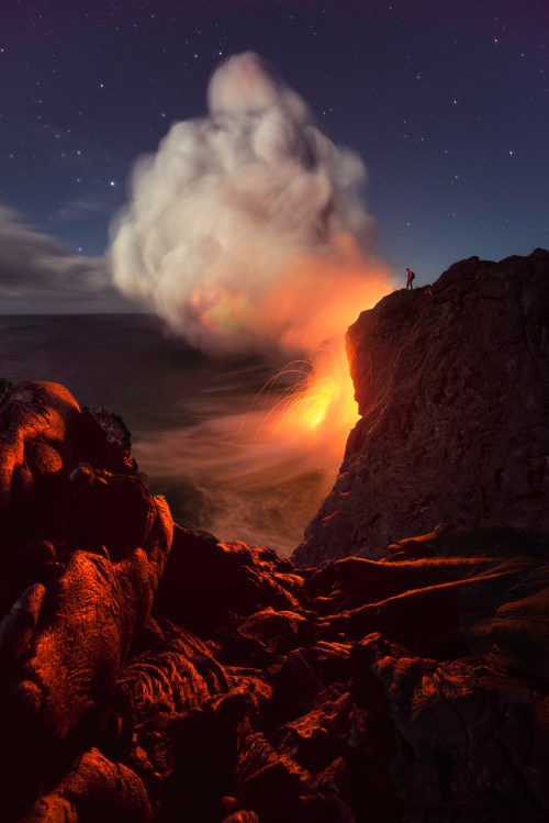 A person stands on a rocky cliff, observing a volcanic eruption under a starry night sky, with glowing lava and smoke.