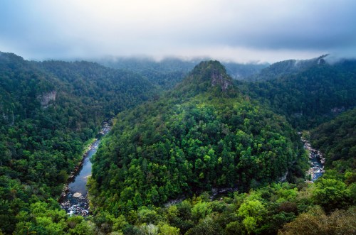 A lush, green valley with a winding river, surrounded by misty mountains and dense forest under a cloudy sky.