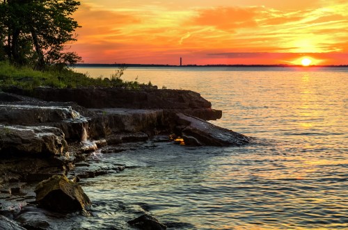 Sunset over a calm lake, with rocky shoreline and trees silhouetted against vibrant orange and yellow sky.