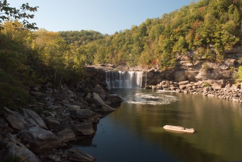 A serene waterfall cascades into a calm lake, surrounded by lush green trees and rocky shores under a clear blue sky.