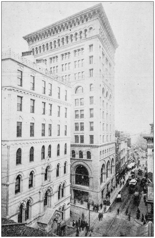 Historic black-and-white photo of a tall building at the intersection of two streets, with people and horse-drawn carriages below.