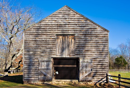 A rustic wooden barn with open doors, set against a clear blue sky and surrounded by a grassy area.