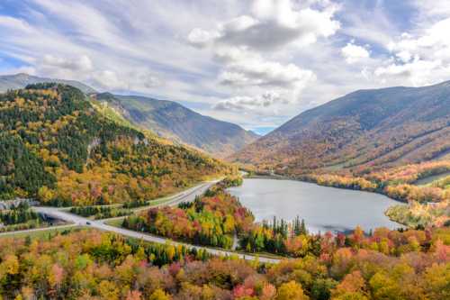 A scenic view of a lake surrounded by colorful autumn foliage and mountains under a partly cloudy sky.