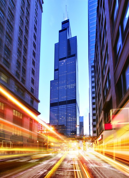 A tall skyscraper towers over a city street, with light trails from passing vehicles creating a dynamic scene at dusk.