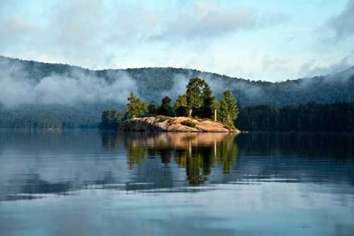 A serene lake scene featuring a small island surrounded by trees, with misty mountains in the background and calm water reflections.