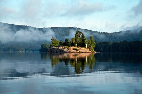 A serene lake scene featuring a small island surrounded by trees, with misty mountains in the background and calm water reflections.