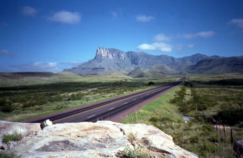 A long, empty road stretches toward a distant mountain under a clear blue sky, surrounded by green fields.