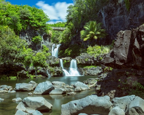 A serene waterfall cascades into a calm pool, surrounded by lush greenery and rocky terrain under a blue sky.