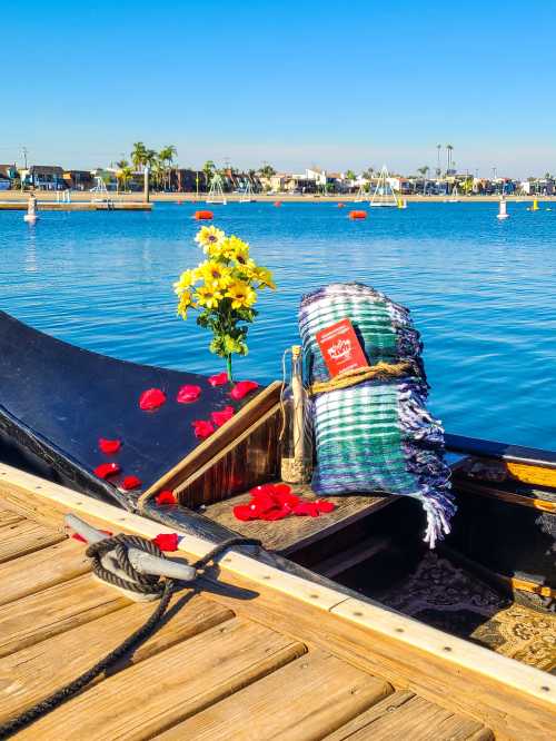 A decorated gondola with flowers, a blanket, and rose petals by a calm blue waterway under a clear sky.