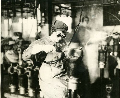 A woman in goggles welds metal in a factory, surrounded by machinery and smoke.