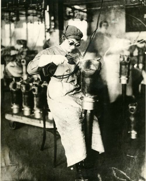 A woman wearing goggles welds a metal piece in a workshop, surrounded by machinery and tools.