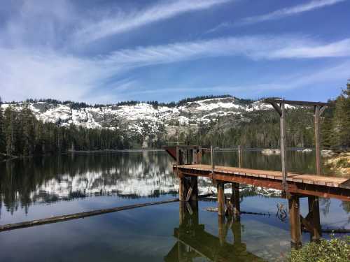 A serene lake with a wooden dock, surrounded by snow-capped mountains and lush green trees under a blue sky.