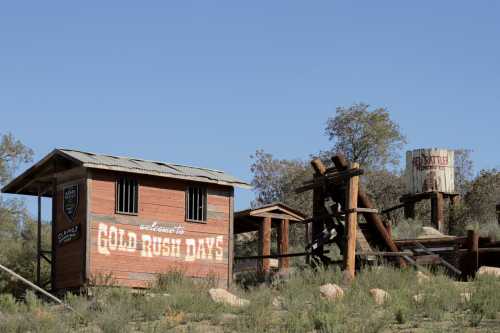 A rustic wooden building with a sign reading "Gold Rush Days" next to a water tower in a grassy area.