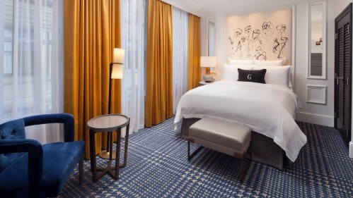 A stylish hotel room featuring a large bed, blue armchair, and elegant curtains, with modern decor and artwork.