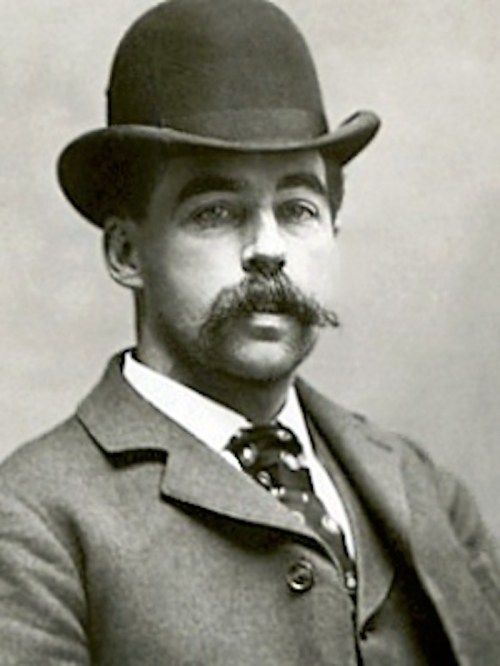 A black-and-white portrait of a man with a mustache, wearing a bowler hat and a suit with a patterned tie.