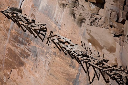 A weathered wooden bridge precariously attached to a rocky cliff face, surrounded by rugged terrain.