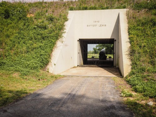 Entrance to Battery Lewis, a concrete structure with a barred gate, surrounded by greenery and a gravel path.