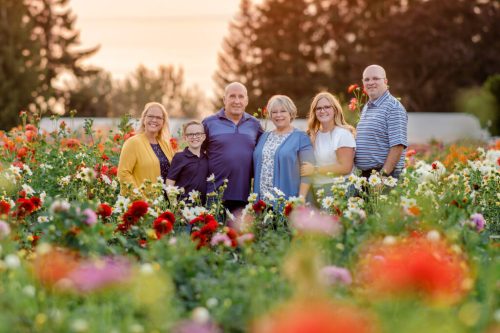 A family stands together in a colorful flower field during sunset, smiling and enjoying the moment.