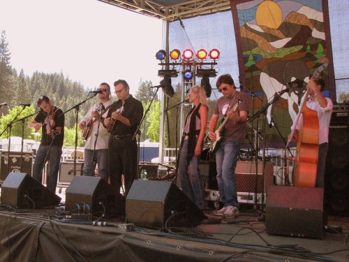 A band performs on stage with instruments, set against a backdrop of trees and a colorful tapestry.