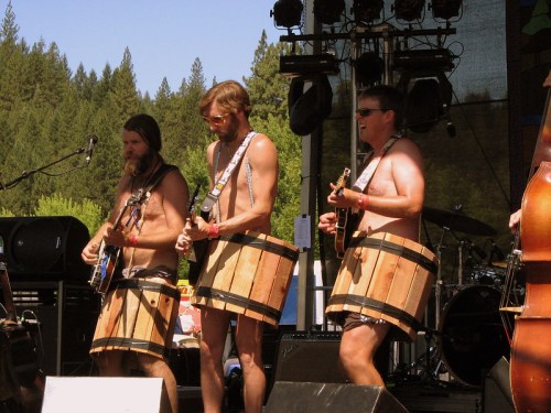Three musicians perform on stage, wearing wooden barrels as costumes, with guitars and a lively backdrop of trees.
