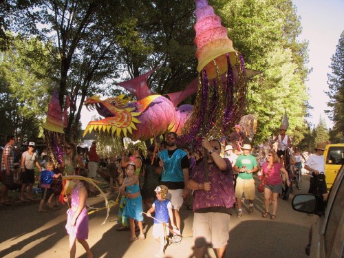 A colorful dragon float leads a lively parade with people in festive attire celebrating among trees.