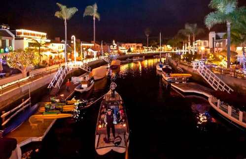 A scenic nighttime view of a canal lined with illuminated buildings and boats, surrounded by palm trees.